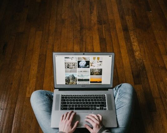 Młody chłopak, który siedzi na drewnianej podłodze z laptopem na kolanach i pracuje jako freelancer.