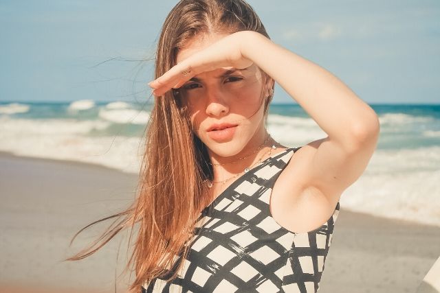 Młoda dziewczyna, z długimi włosami zasłania oczy przed słońcem. Stoi na plaży i ma na sobie kostium kąpielowy w biało-czarny geometryczny wzór. Opalanie jest skutkiem, jaki wywołuje promieniowanie UV.