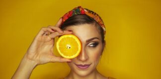 Czy nadmiar witaminy C w organizmie w ogóle jest możliwy? Na zdjęciu młoda dziewczyna, która plastrem pomarańczy zasłania jedno oko. Stoi na tle żółtej ściany.