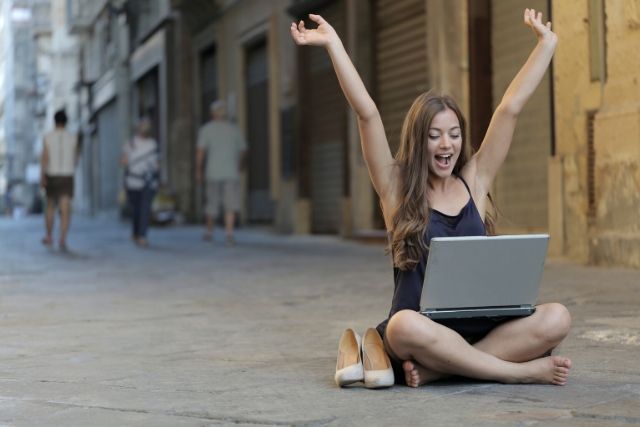 Młoda dziewczyna, która siedzi boso na deptaku z komputerem na kolanach. Obok niej stoją jej szpilki. Ma wyciągnięte do góry ręce i jest usmiechnięta. Produktywność jest kluczem do sukcesu zawodowego.