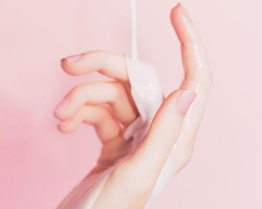 Kobieca dłoń na różowym tle, na którą leje się emulsja nawilżająca. Grafika ilustrująca problem, jakim są suche dłonie.