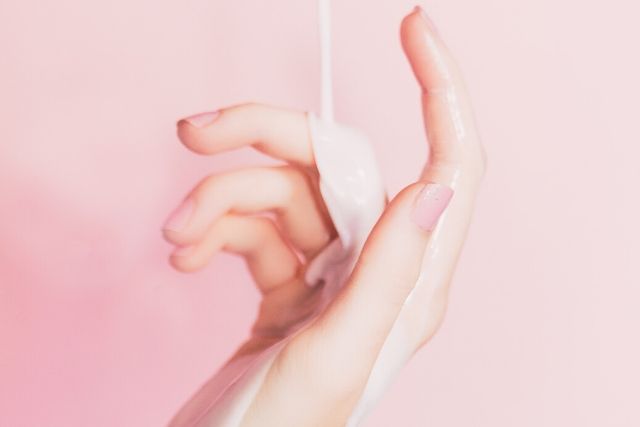 Kobieca dłoń na różowym tle, na którą leje się emulsja nawilżająca. Grafika ilustrująca problem, jakim są suche dłonie.
