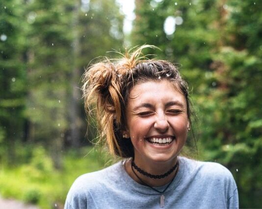 Uśmiechnięta młoda dziewczyna odczuwająca szczęście. Ma spięte w kok włosy, a za jej plecami widać las.