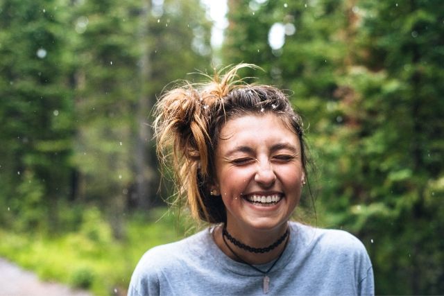 Uśmiechnięta młoda dziewczyna odczuwająca szczęście. Ma spięte w kok włosy, a za jej plecami widać las.